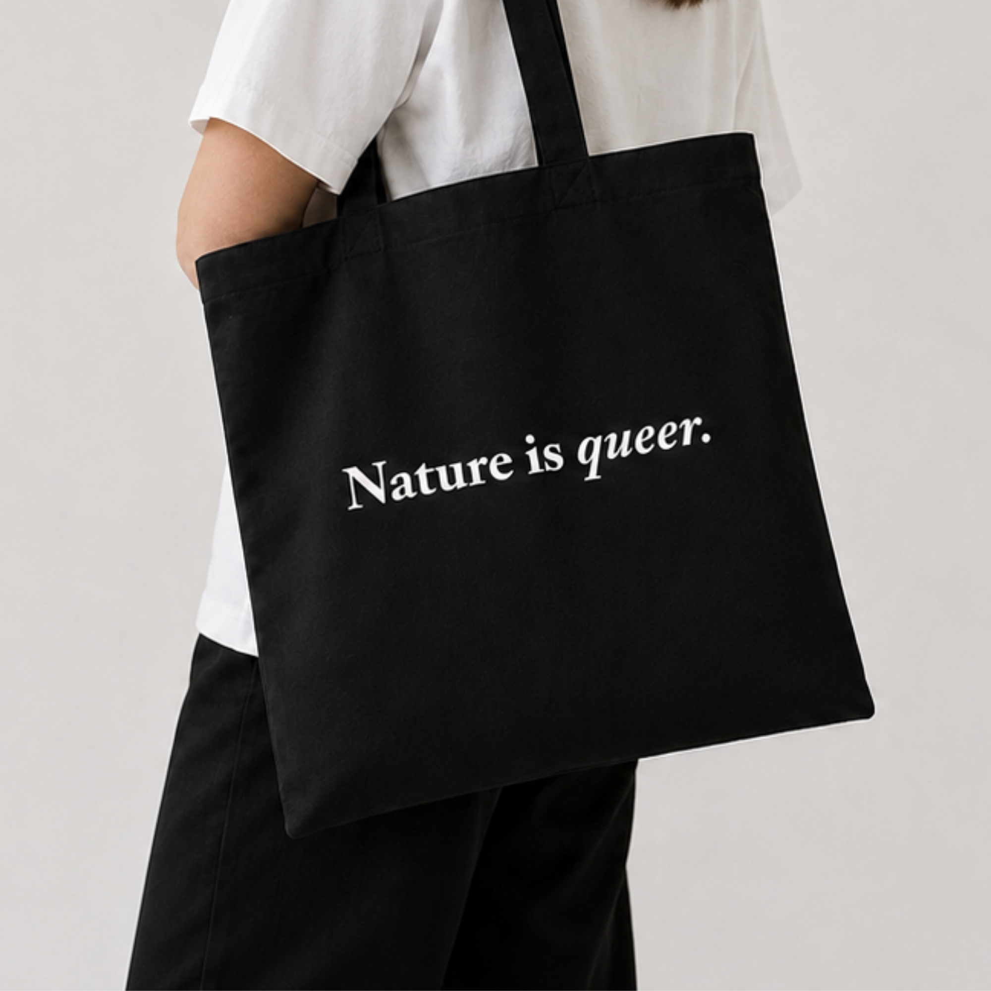 A person wearing a white t-shirt and black pants carries a black canvas tote bag with the white printed text 'Nature is queer.' on the side, photographed against a light gray studio background.