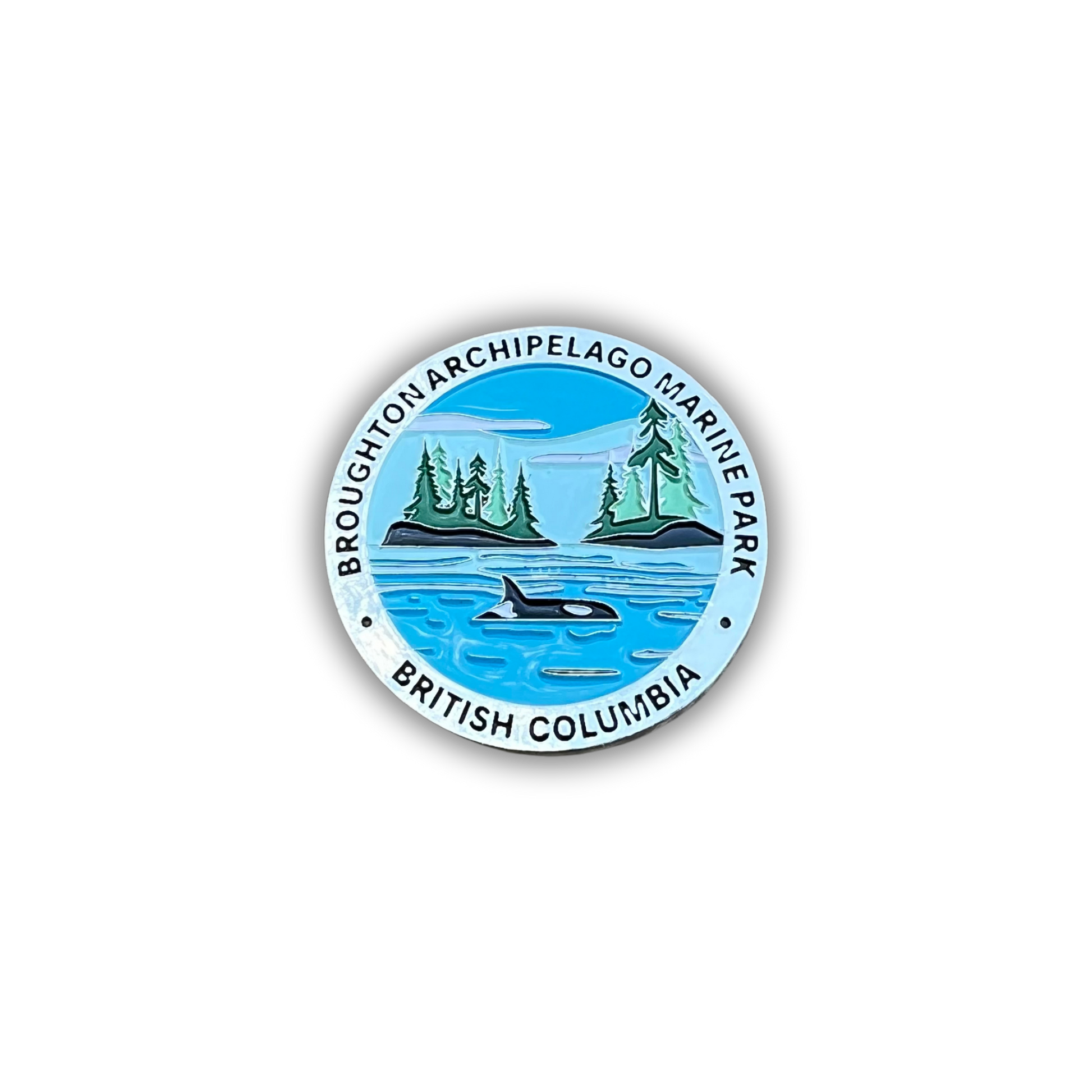 A circular pin badge featuring the Broughton Archipelago Marine Park in British Columbia, Canada. The design shows a blue ocean with an orca whale, two green islands with pine trees, and a light blue sky, all enclosed by a white border with black text reading 'BROUGHTON ARCHIPELAGO MARINE PARK' and 'BRITISH COLUMBIA'.