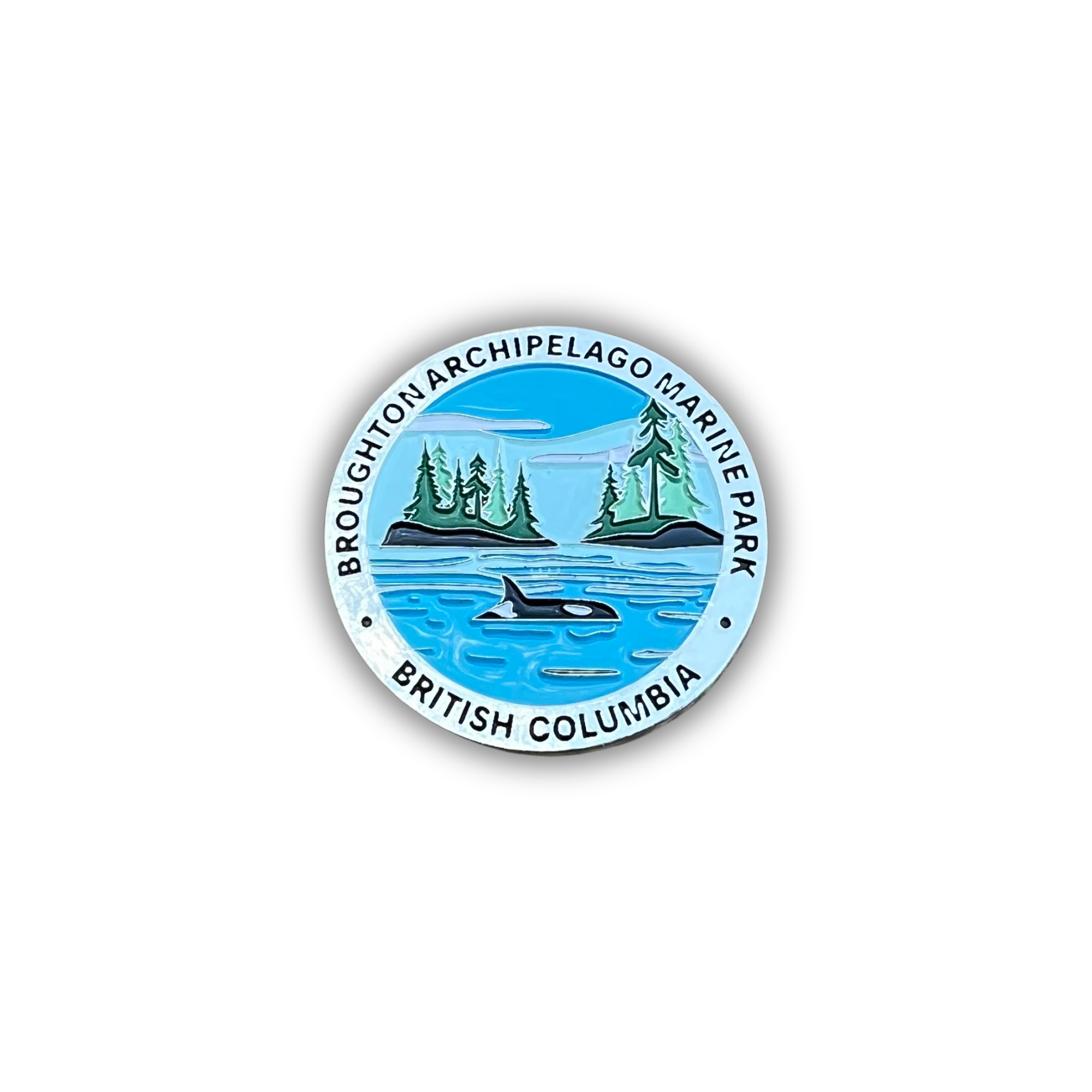 A circular pin badge featuring the Broughton Archipelago Marine Park in British Columbia, Canada. The design shows a blue ocean with an orca whale, two green islands with pine trees, and a light blue sky, all enclosed by a white border with black text reading 'BROUGHTON ARCHIPELAGO MARINE PARK' and 'BRITISH COLUMBIA'.