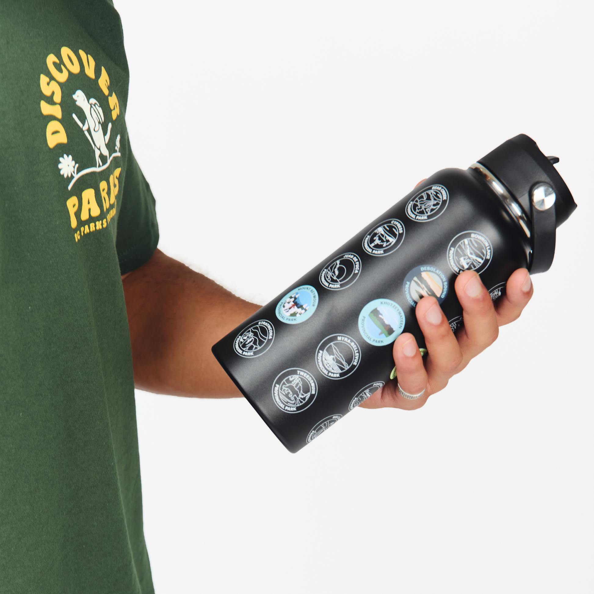 A person in a green BC Parks t-shirt holds a black insulated BC Parks Sticker water bottle covered in circular travel-style stickers, against a plain light gray background.