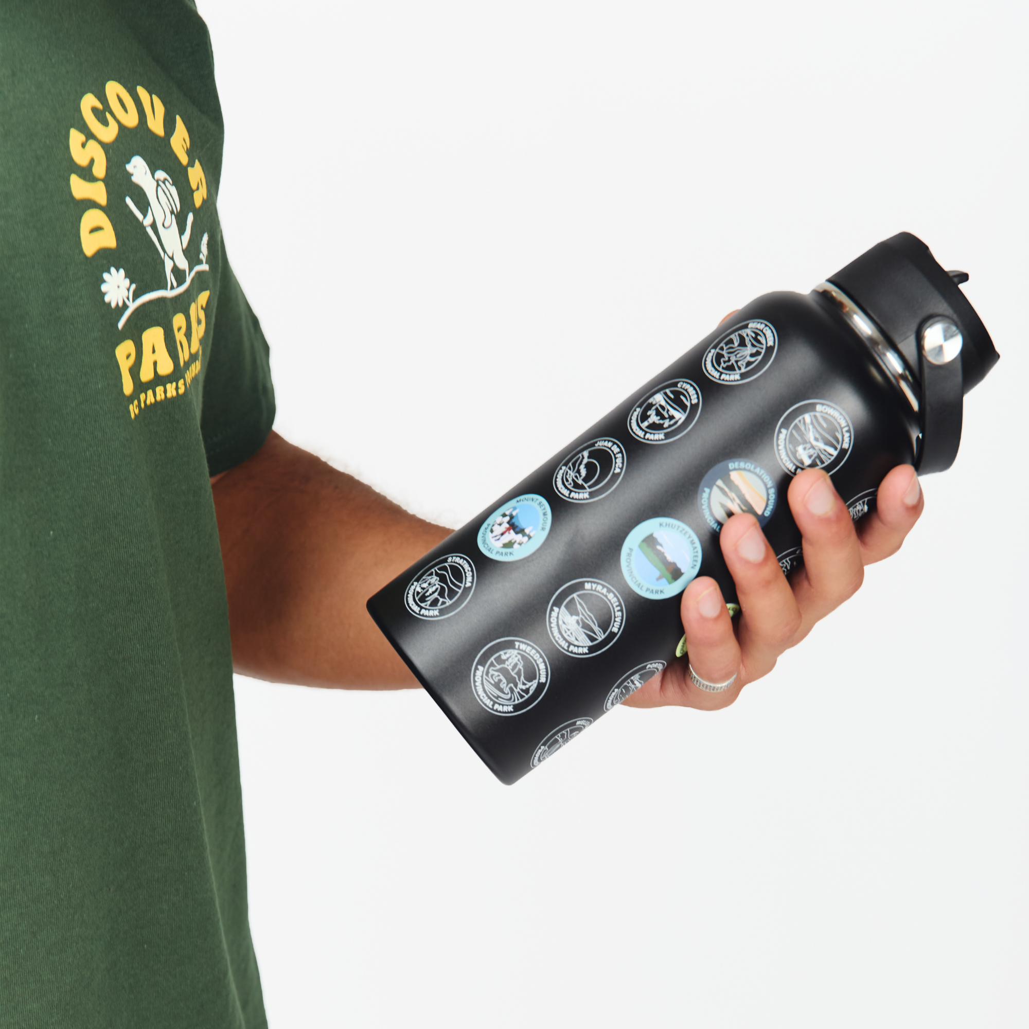 A person in a green BC Parks t-shirt holds a black insulated BC Parks Sticker water bottle covered in circular travel-style stickers, against a plain light gray background.