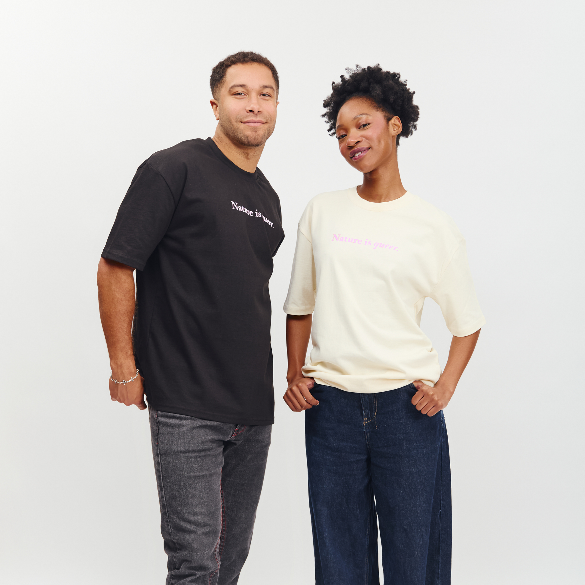 A man and a woman modeling oversized graphic t-shirts against a plain light gray studio background. The man wears a black t-shirt with white text, and the woman wears a cream-colored t-shirt with pink text.