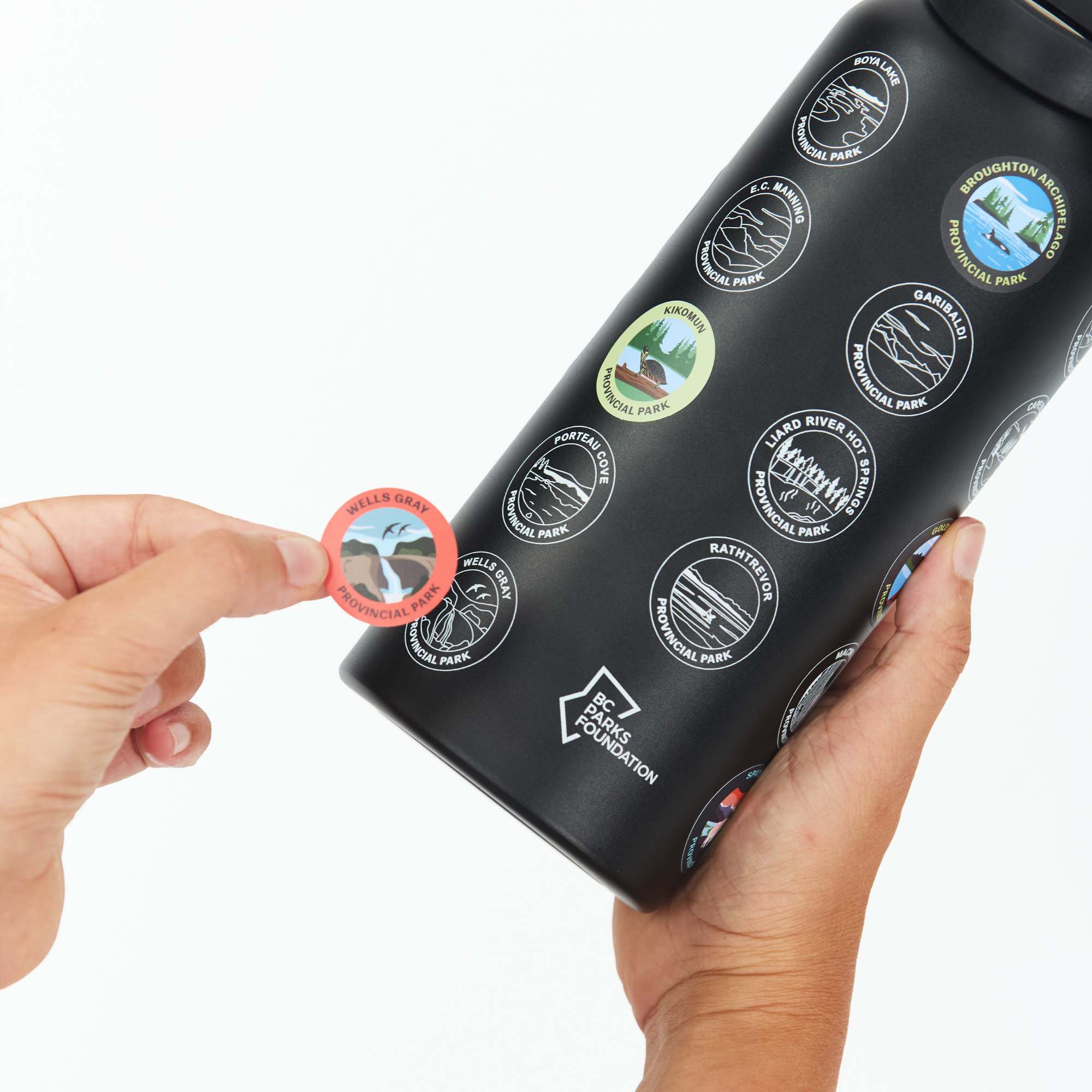 A black reusable water bottle held in a hand, with a second hand placing a round sticker on it. The bottle is covered in circular outdoor-themed stickers, and the bottom shows the Recycled Water Bottle brand logo.