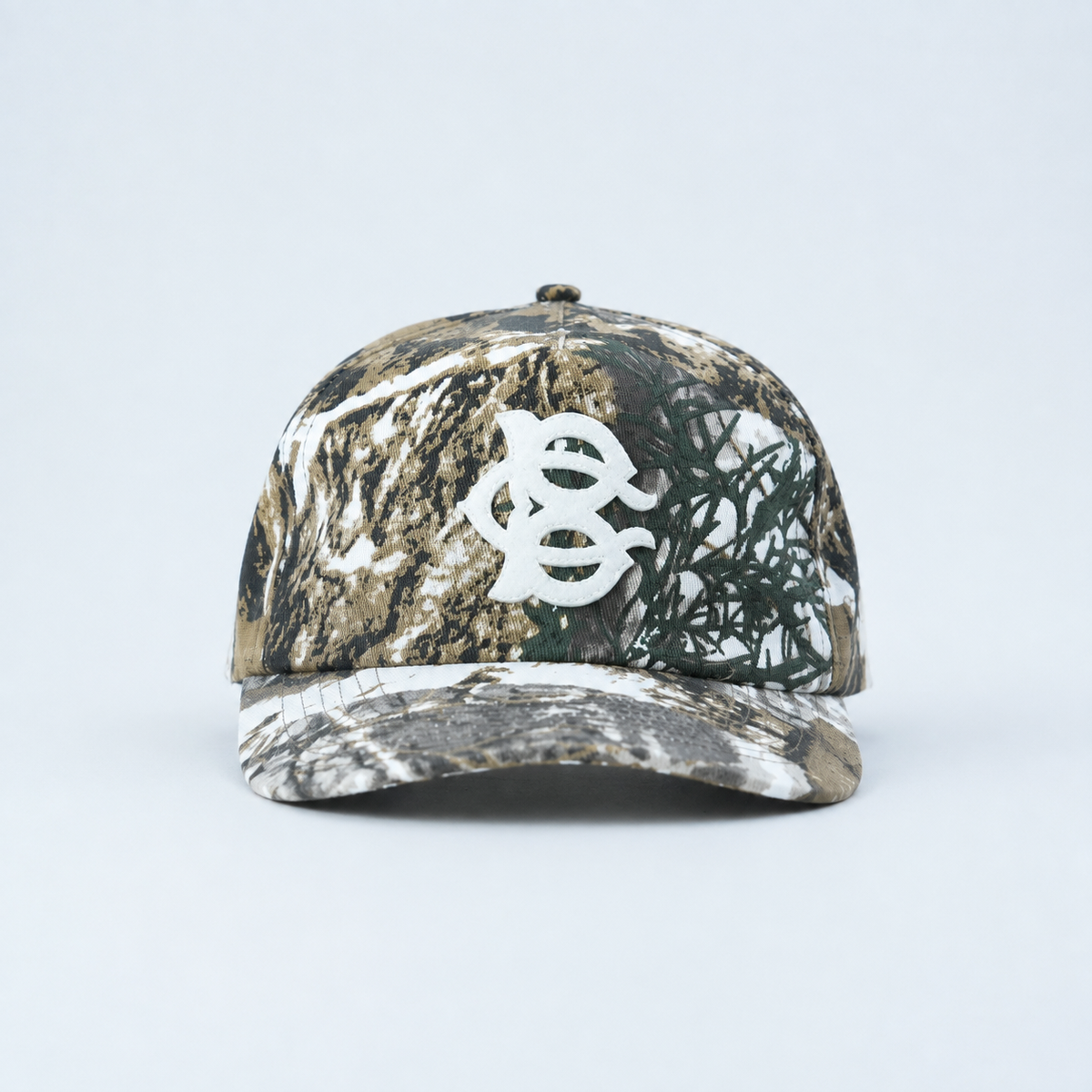 A structured baseball cap featuring  a BC Monogram felt Applique on a realistic woodland camouflage pattern with brown, black, green, and white tones, shown from a side profile on a light gray background.