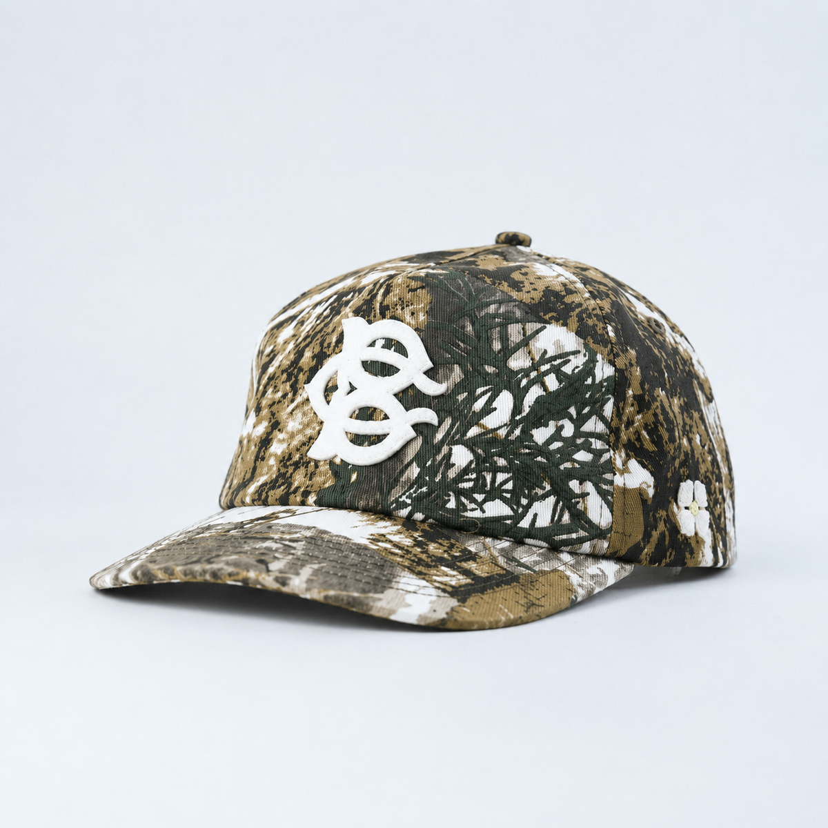 A structured baseball cap featuring  a BC Monogram felt Applique on a realistic woodland camouflage pattern with brown, black, green, and white tones, shown from a side profile on a light gray background.