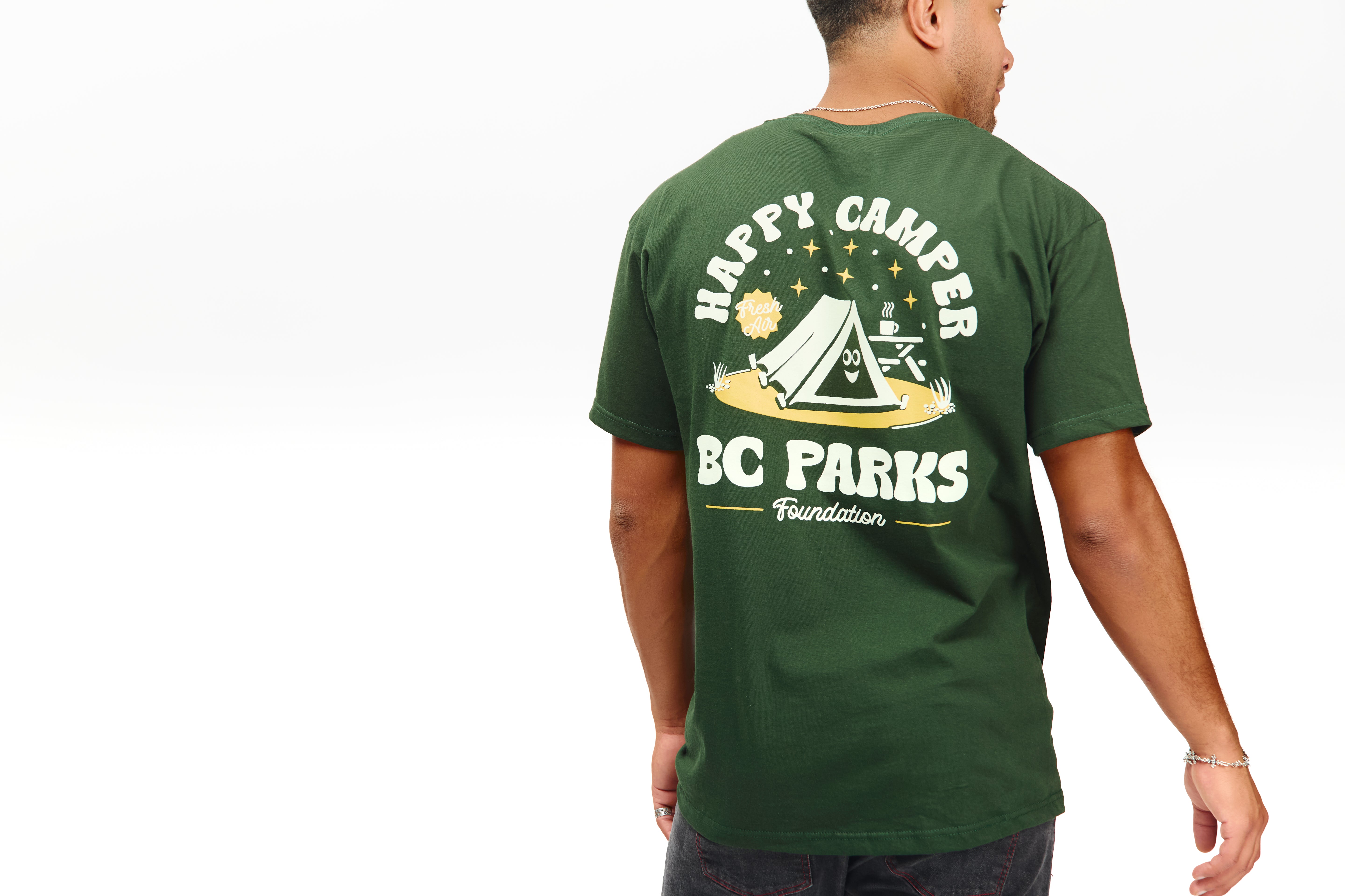 A man shown from the back wearing a dark green t-shirt with a large white and yellow graphic on the back that reads 'HAPPY CAMPER BC PARKS Foundation' with a tent illustration.
