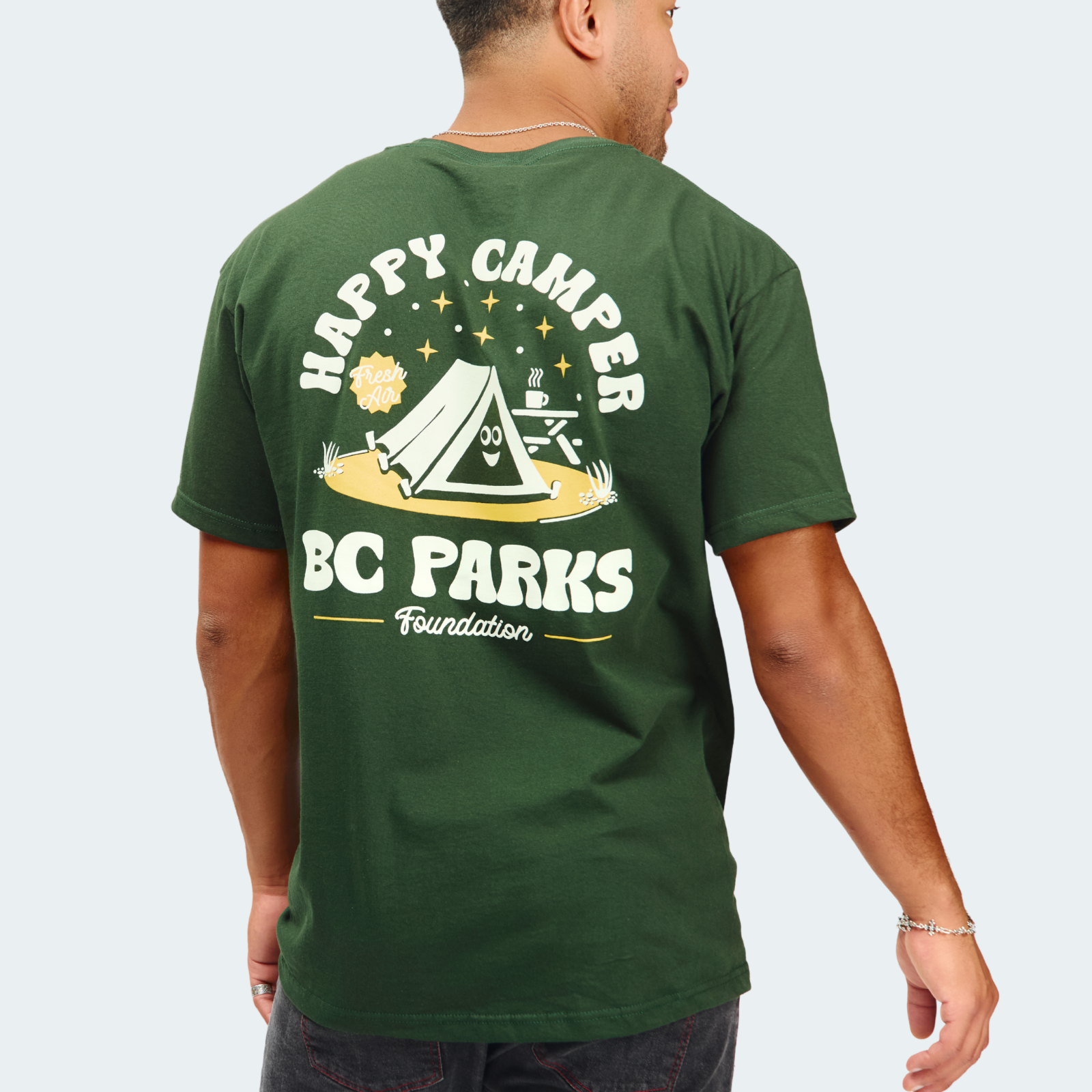 A man wearing a dark green t-shirt, viewed from the back. The shirt features a large graphic print on the back with a camping tent, stars, a sun, and the text 'HAPPY CAMPER BC PARKS Foundation' in white and yellow.