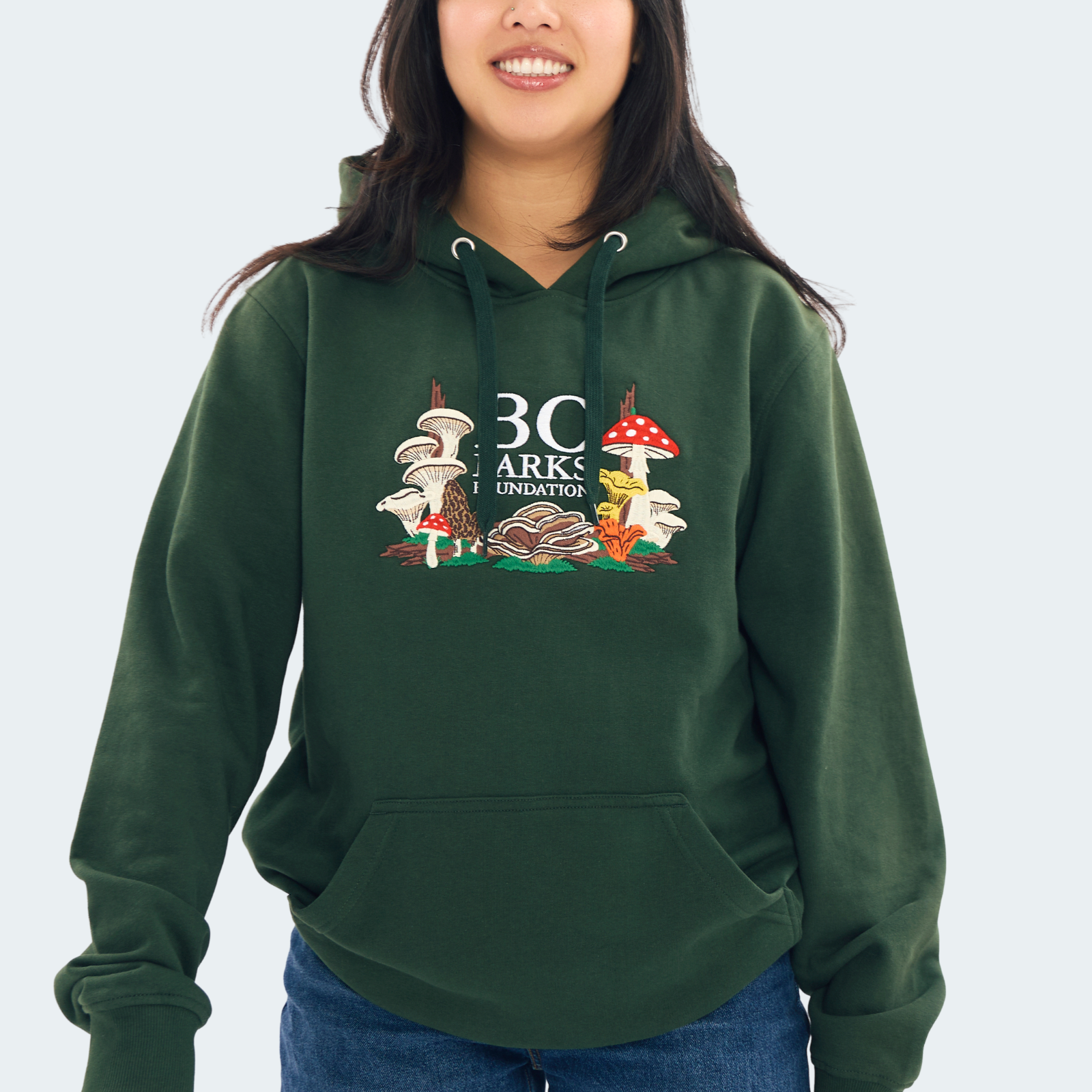 A woman wearing a Forest green pullover hoodie with a colourful graphic on the chest featuring mushrooms and the text 'BC PARKS FOUNDATION'.