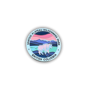 A circular enamel pin featuring a stylized landscape of Spatsizi Plateau Wilderness Park in British Columbia, with a light blue background, pink and dark blue mountains, and a white caribou. The text 'SPATSIZI PLATEAU WILDERNESS PARK' and 'BRITISH COLUMBIA' is printed around the border.