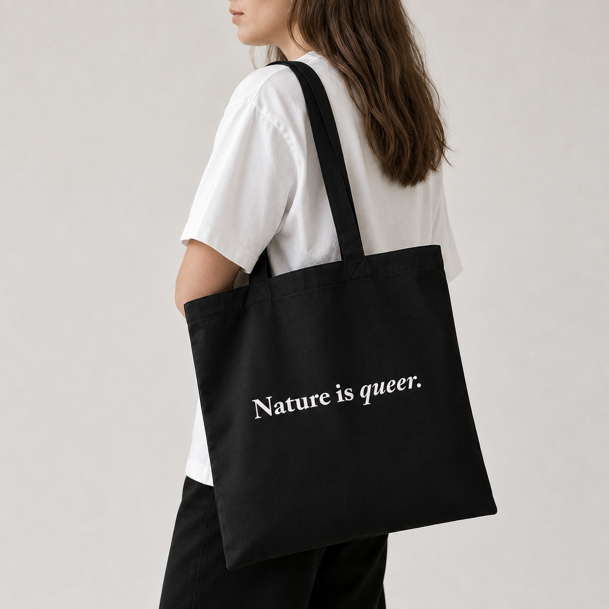 A person wearing a white t-shirt and black pants carries a black canvas tote bag with the white printed text 'Nature is queer.' on the side, photographed against a light gray studio background.
