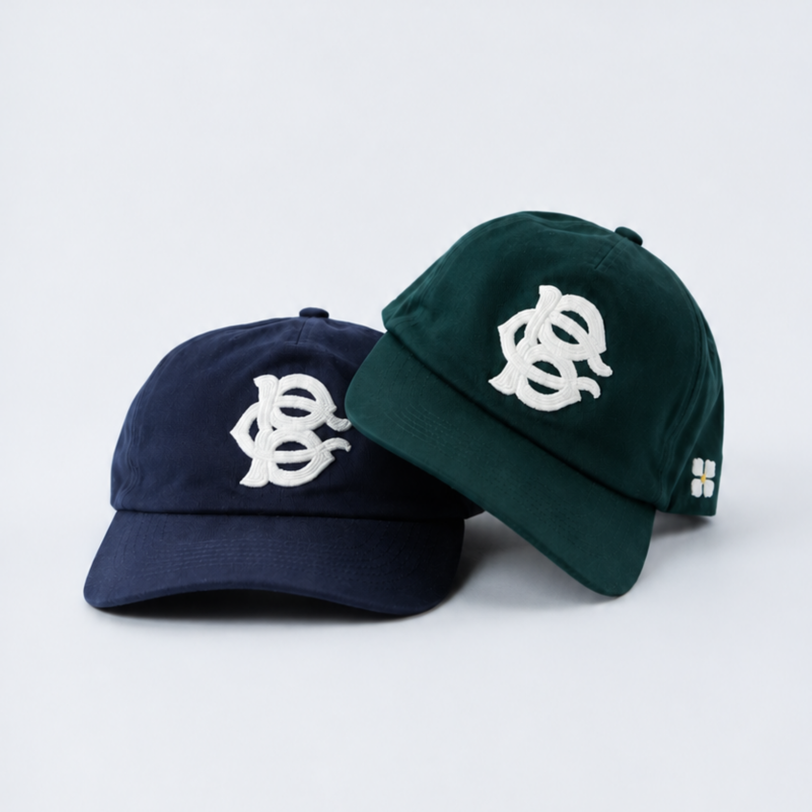 Two BC Monogram baseball caps on a light gray studio background: one navy blue and one forest green, each with a large white embroidered stylized logo on the front and a small white embroidered patch on the side.