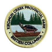 A circular embroidered patch for Kikomun Creek Provincial Park, British Columbia, featuring a painted turtle on a log by a lake with pine trees in the background, surrounded by black embroidered text on a cream border.